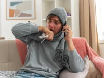 Sick young man with scarf around neck wearing winter hat holding his hand close mouth talking phone sitting couch living room scaled 1 215x161
