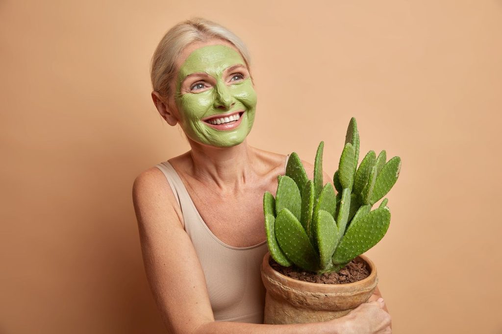 Positive good looking european woman senior woman applies green beauty mask holds cactus pot dressed casual wear isolated beige wall 1024x682